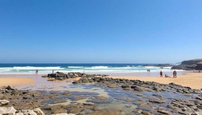 A sunny beach with clear waves, golden sands perfect for sunbathing, rocky outcrops for exploration, accessible ramps, and diverse people enjoying the day under a blue sky.